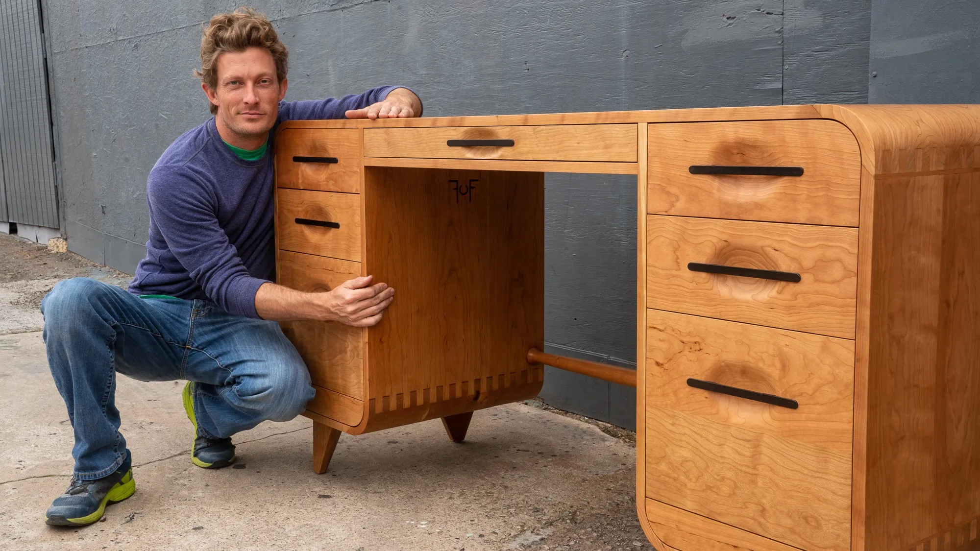 Bryan Ferris crouching beside the finished cherry executive desk, showing its full width, two drawer banks and open knee space.