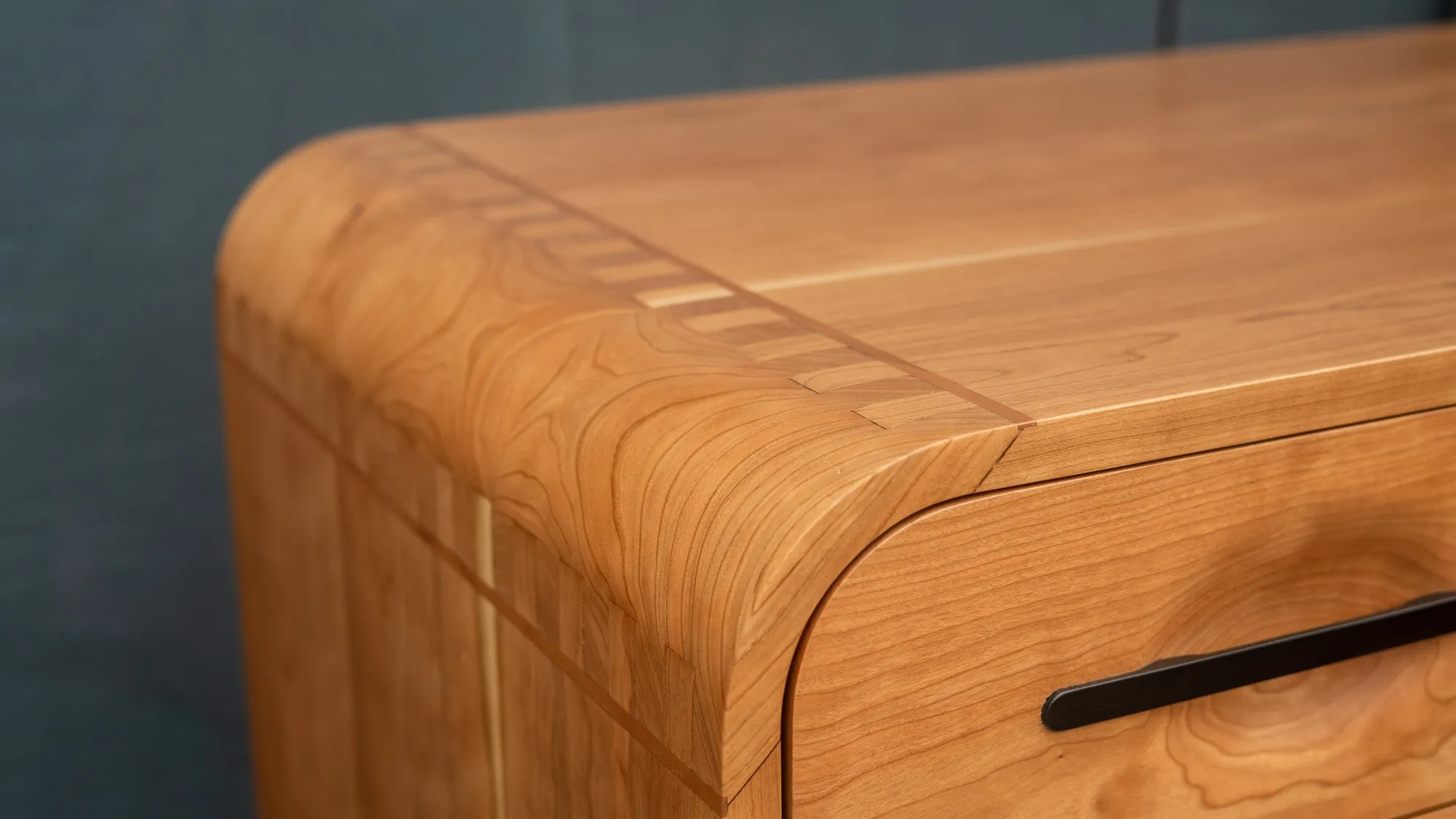 The front corner of the cherry desk, where the top rolls over and the finger-jointed corner is picked out by a fine light line.
