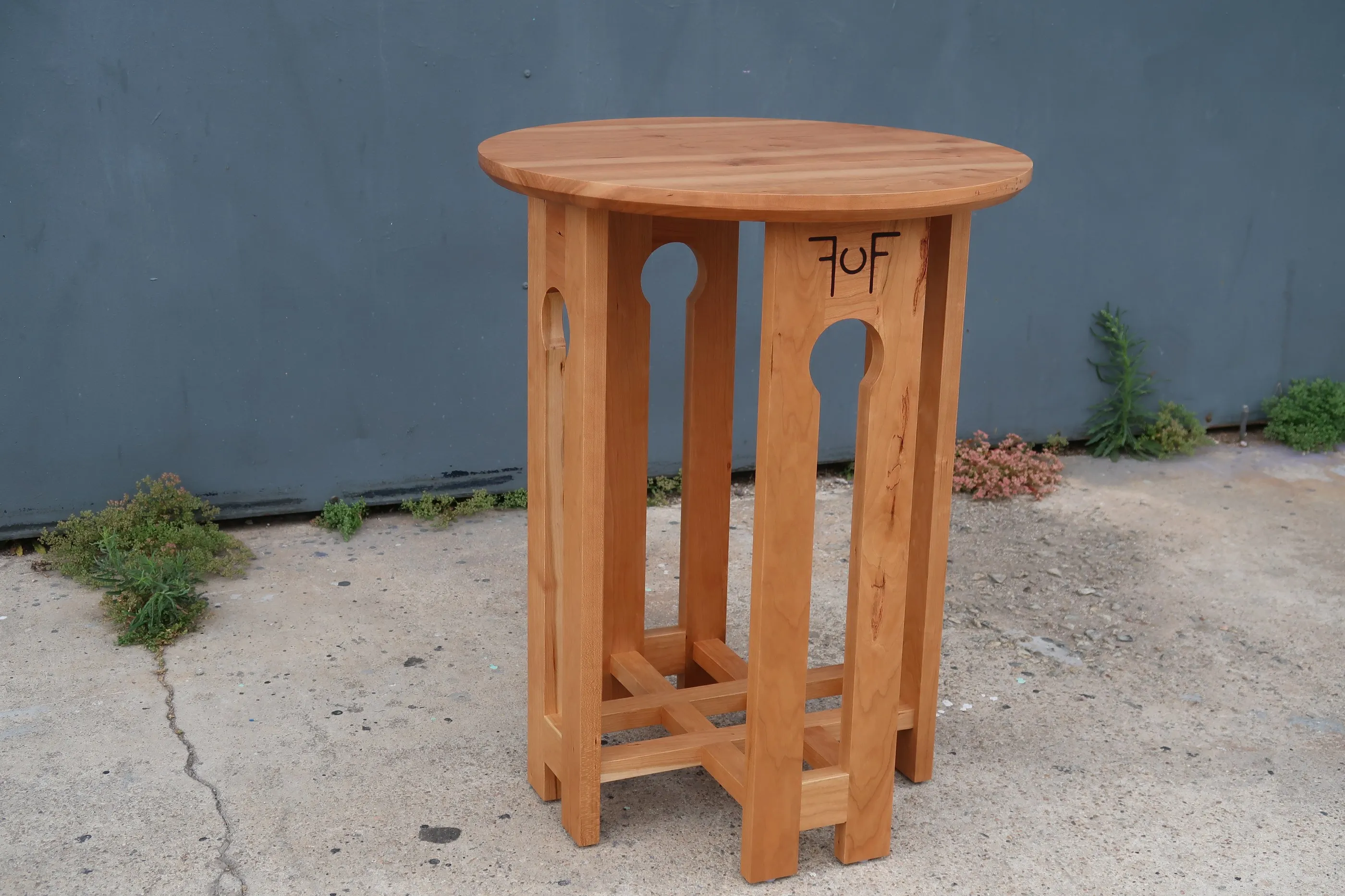 The craftsman end table in cherry, seen at an angle against a slate-painted wall, with its round top and the keyway cutouts in the legs visible.