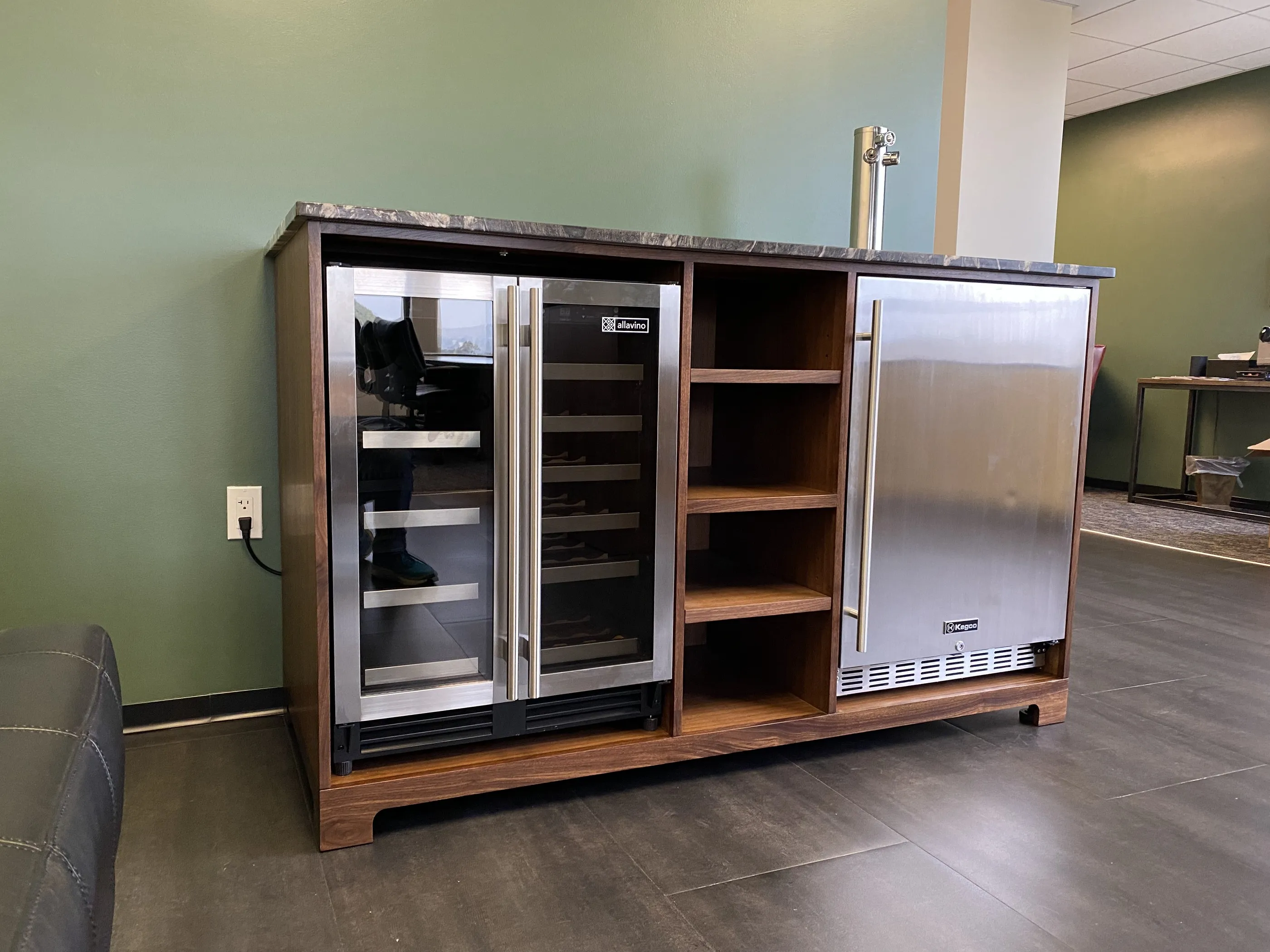 The walnut bar cabinet seen at an angle, housing a stainless wine refrigerator, open shelving and a kegerator with a chrome tap tower under a stone top.